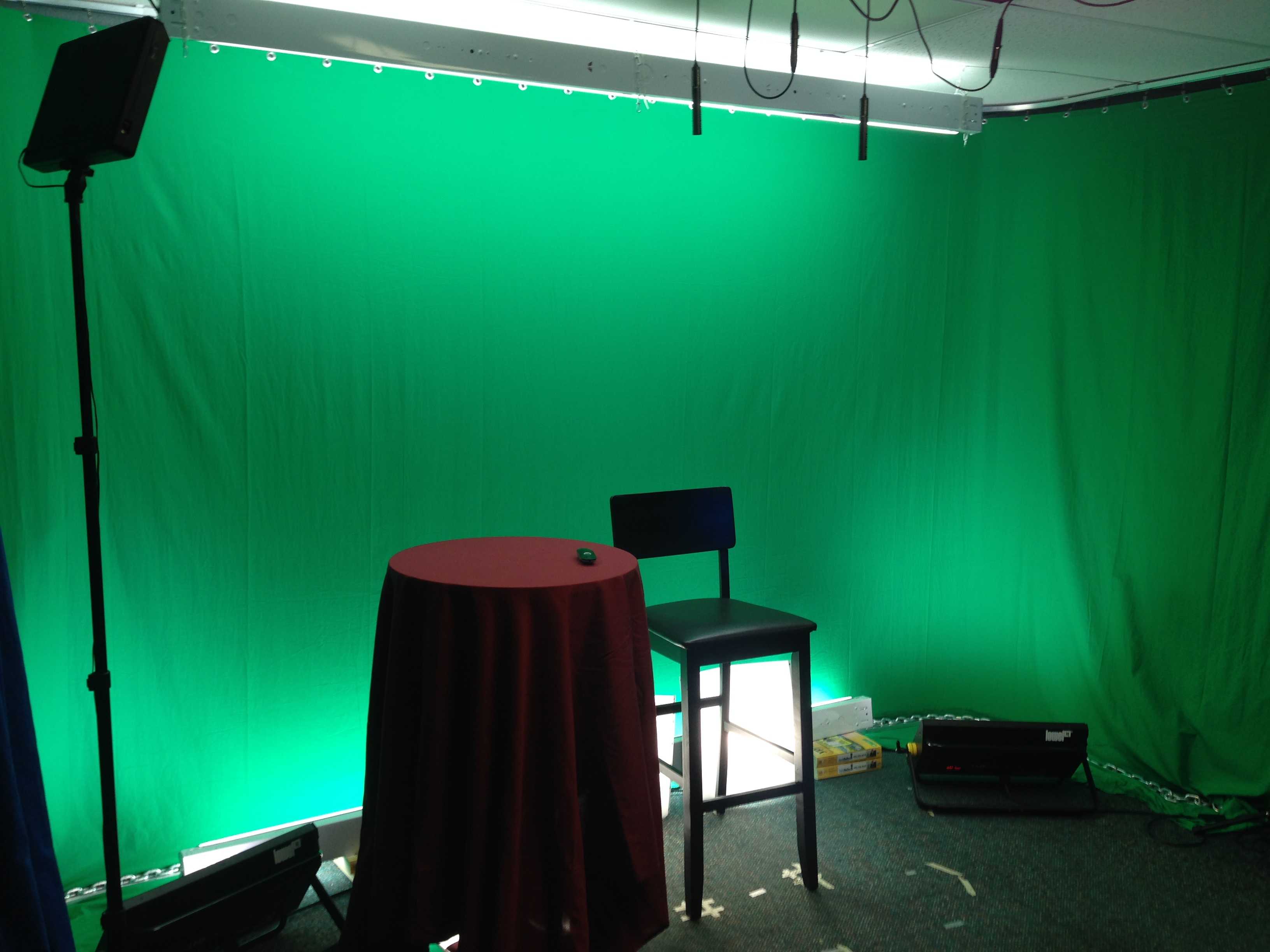 Empty studio space with a green screen backdrop, a small table draped in dark cloth, a stool, and lighting equipment, suggesting a calm and professional training environment